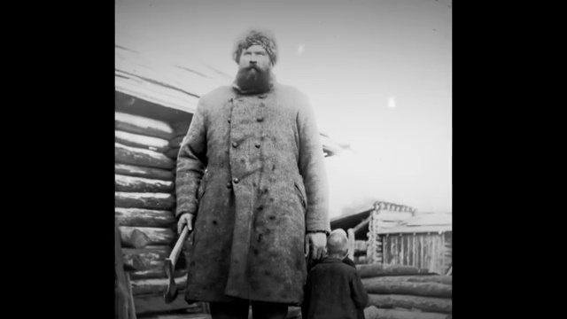 Un homme immense posant près d'une cabane de campement dont la porte et l'architecture sont construites à son échelle.