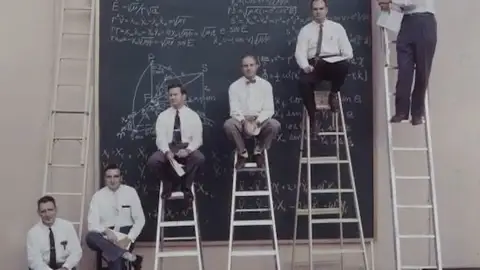 Photographie d'archive montrant des ingénieurs assis sur des échelles devant un tableau noir rempli d'équations complexes.